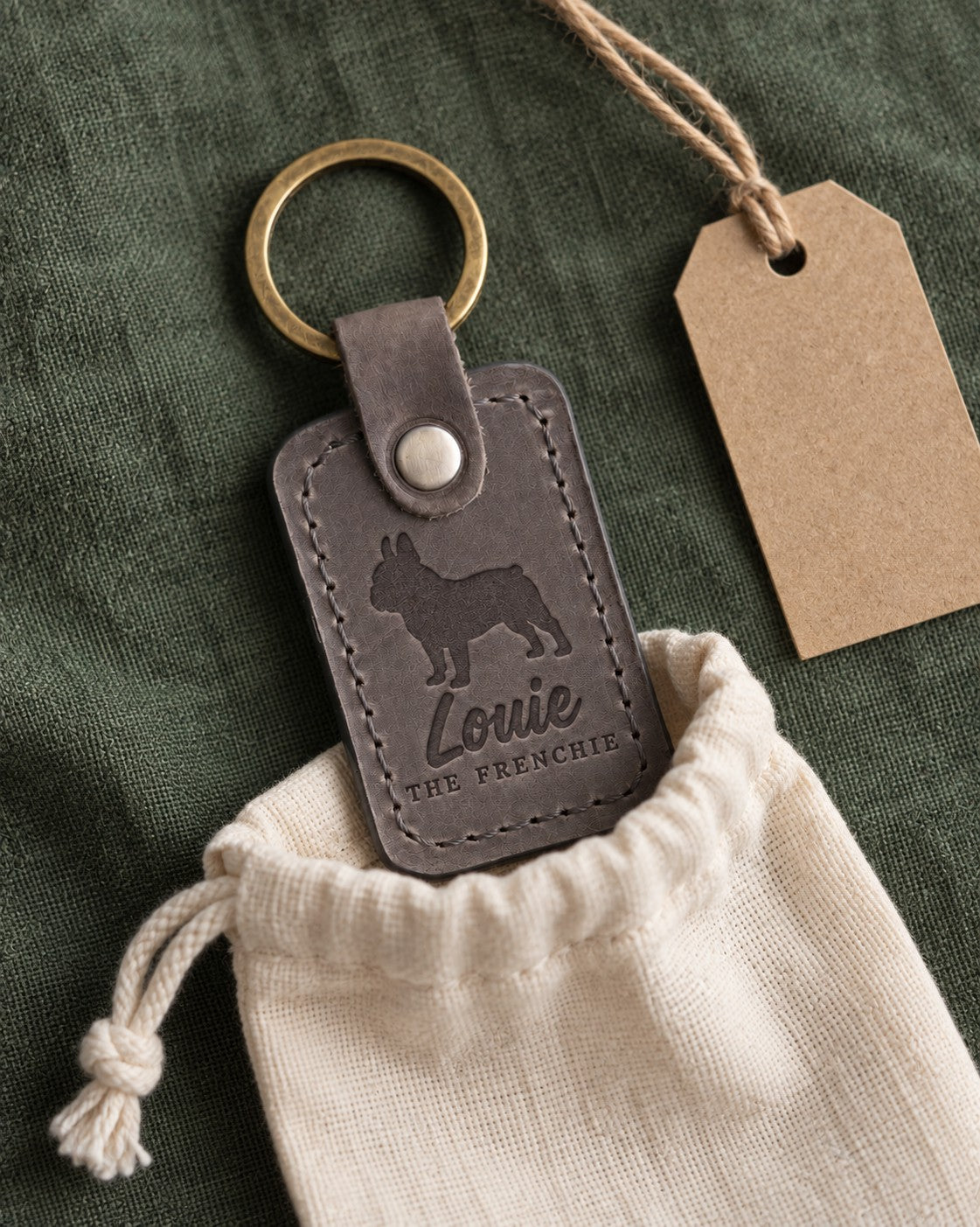 Louie the Frenchie keychain in gray leather tucked into a cotton gift pouch on green linen beside a kraft tag