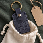 Navy Frenchie Dad keychain half out of a cotton drawstring pouch on dark green linen, a kraft tag beside it