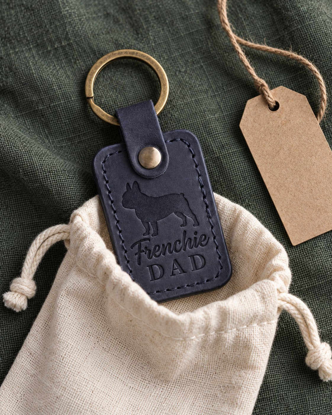 Navy Frenchie Dad keychain half out of a cotton drawstring pouch on dark green linen, a kraft tag beside it
