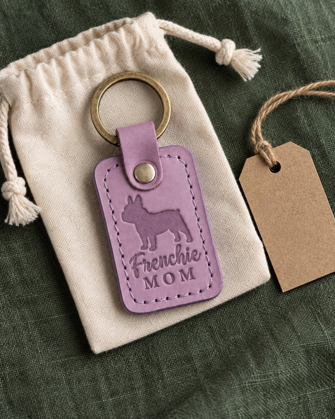 Lilac Frenchie Mom keychain resting on a cotton drawstring pouch on green linen with a kraft gift tag