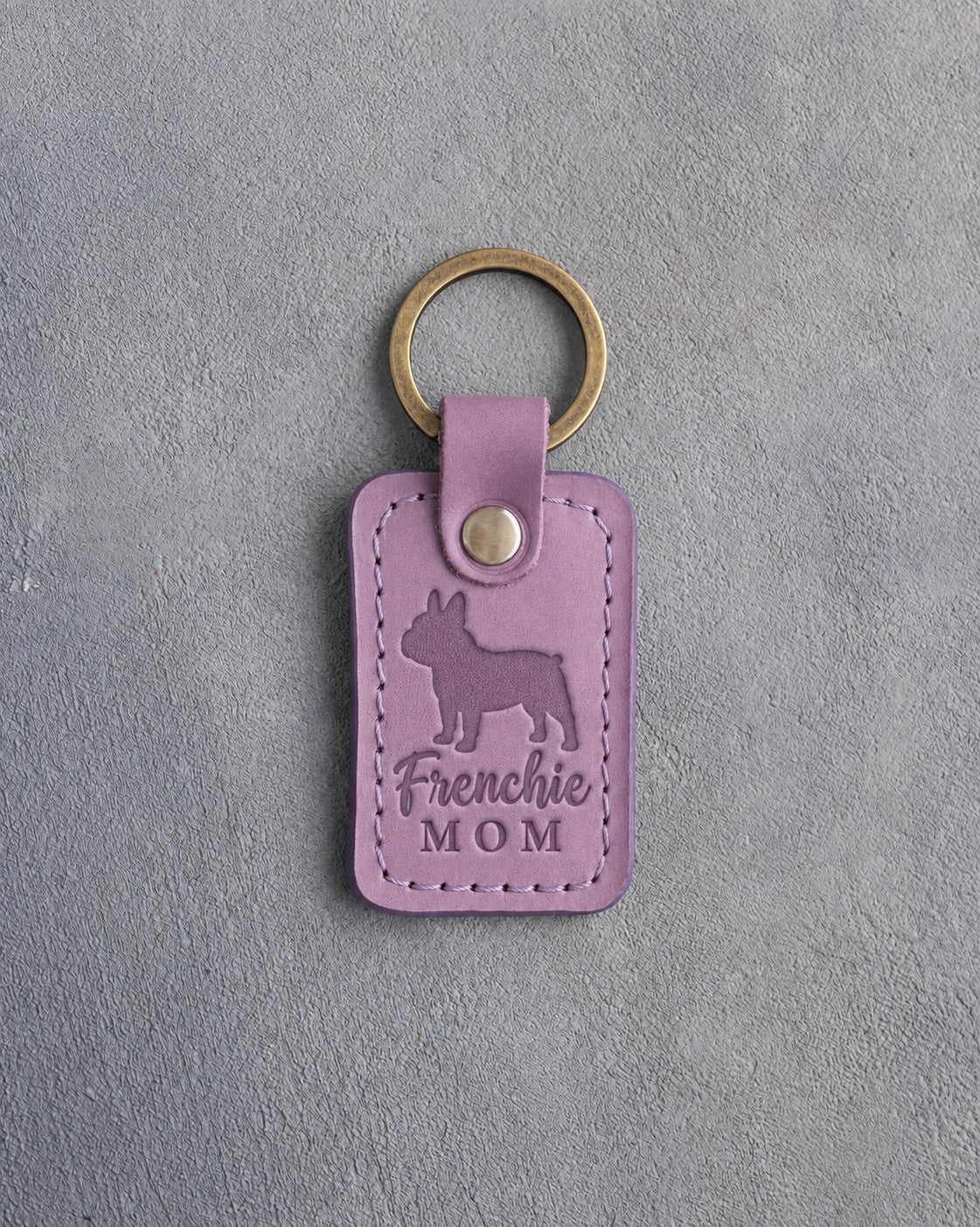 Lilac leather keychain debossed with a French bulldog silhouette and the words Frenchie MOM on gray concrete