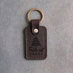 Dark espresso leather keychain debossed with a fir tree above Fresh cut TREES, on gray concrete
