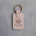 Gray sand leather keychain debossed with an egg basket above the words Fresh eggs DAILY, on gray concrete
