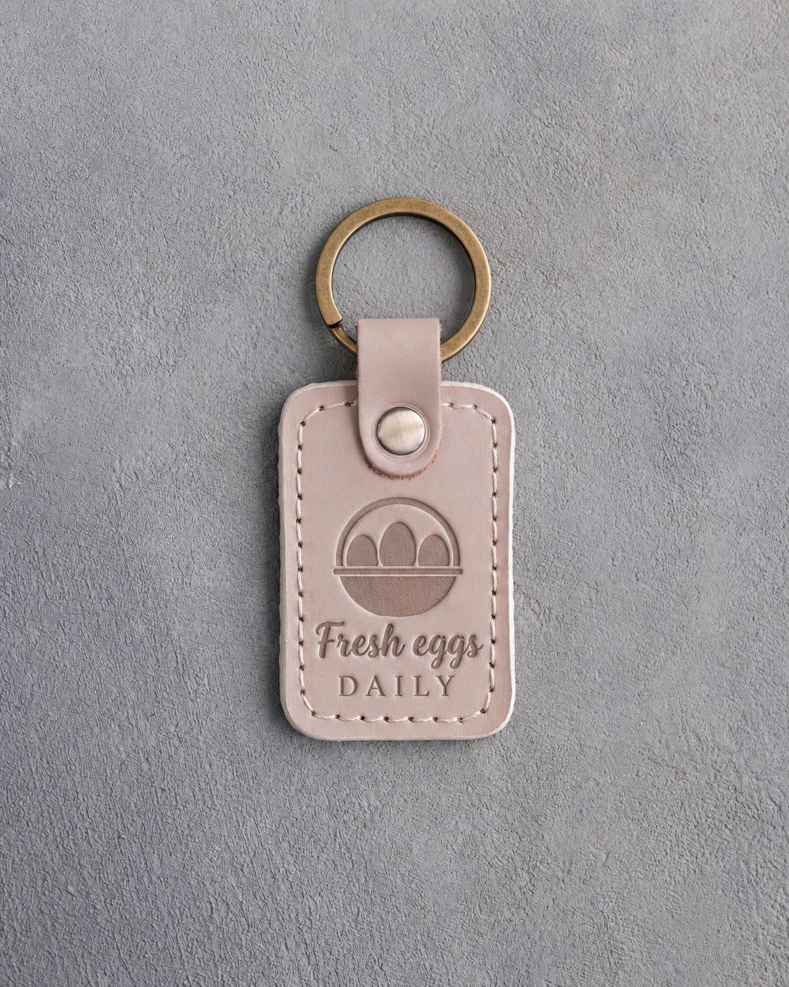 Gray sand leather keychain debossed with an egg basket above the words Fresh eggs DAILY, on gray concrete