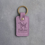 Lilac leather keychain debossed with a golden retriever among three snowflakes and the words Fresh snow ENTHUSIAST, on gray concrete