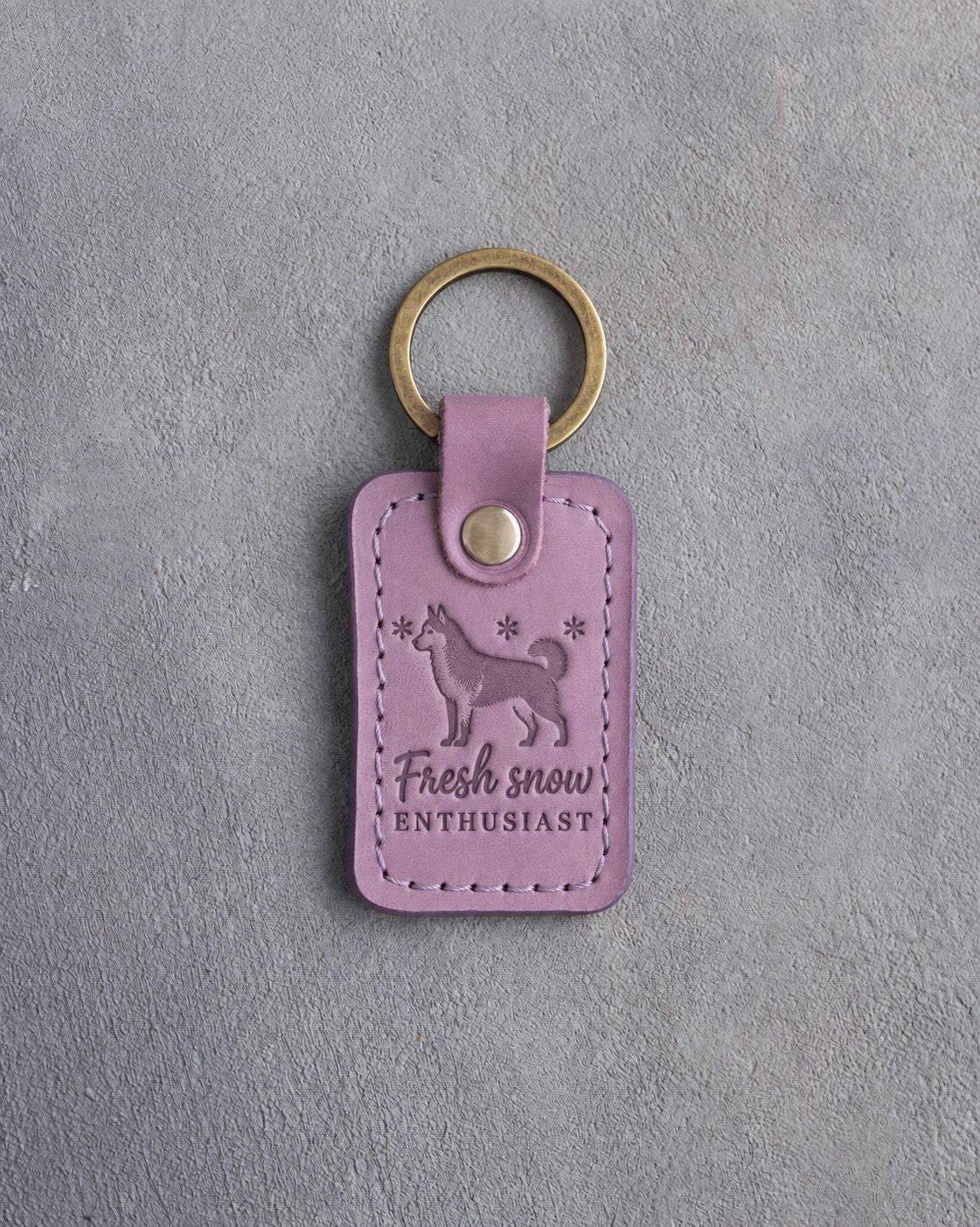 Lilac leather keychain debossed with a golden retriever among three snowflakes and the words Fresh snow ENTHUSIAST, on gray concrete