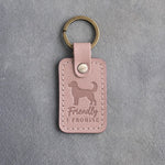 Blush pink leather keychain debossed with an alert standing dog and the words Friendly I PROMISE, on gray concrete