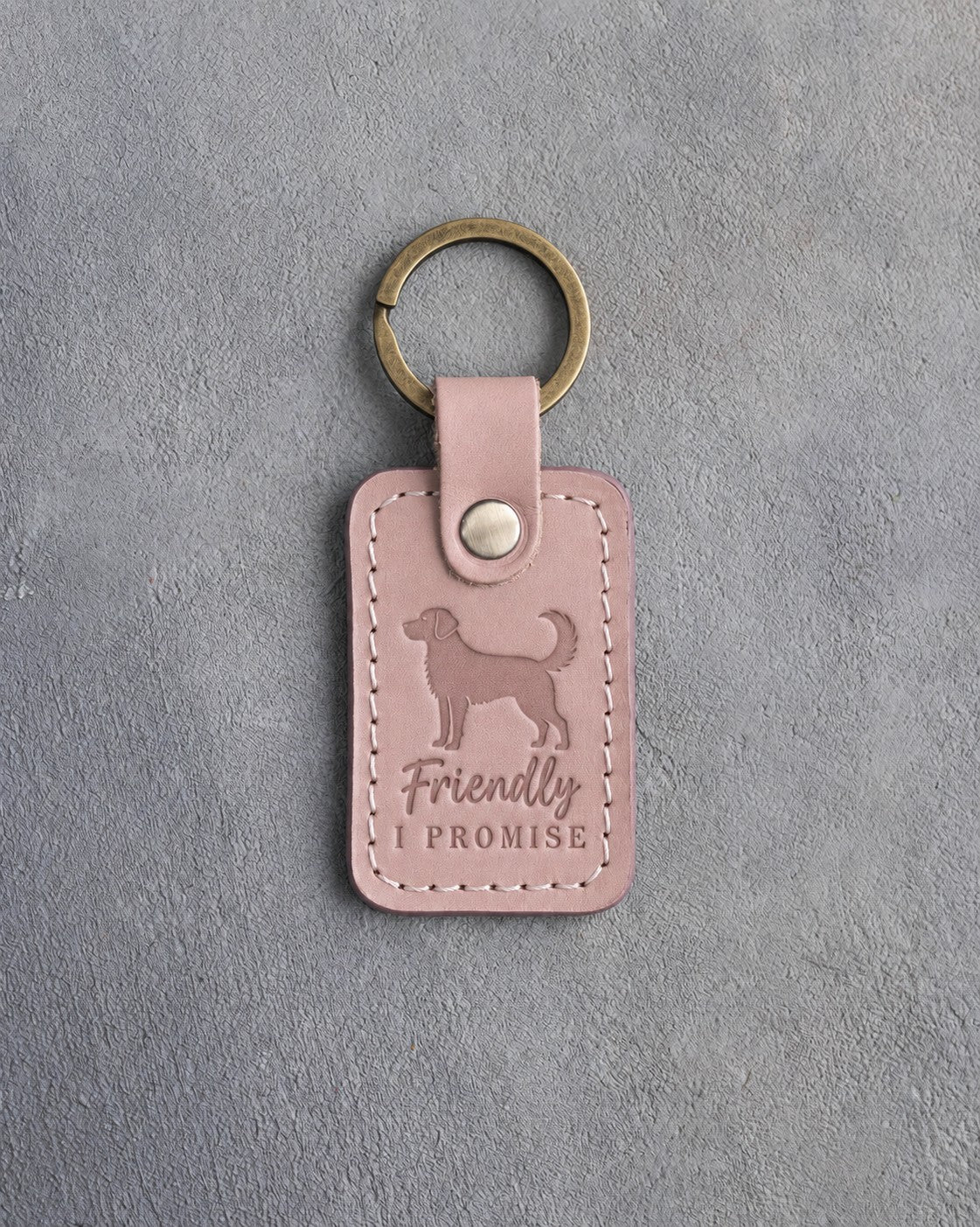 Blush pink leather keychain debossed with an alert standing dog and the words Friendly I PROMISE, on gray concrete