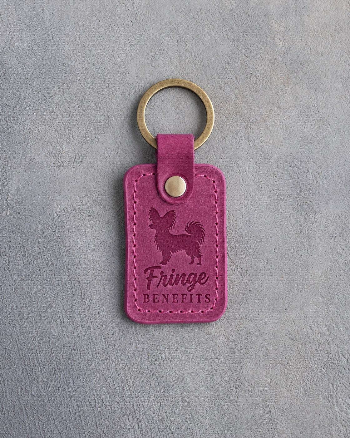Fuchsia pink leather keychain debossed with a Papillon silhouette and the words Fringe BENEFITS, on gray concrete