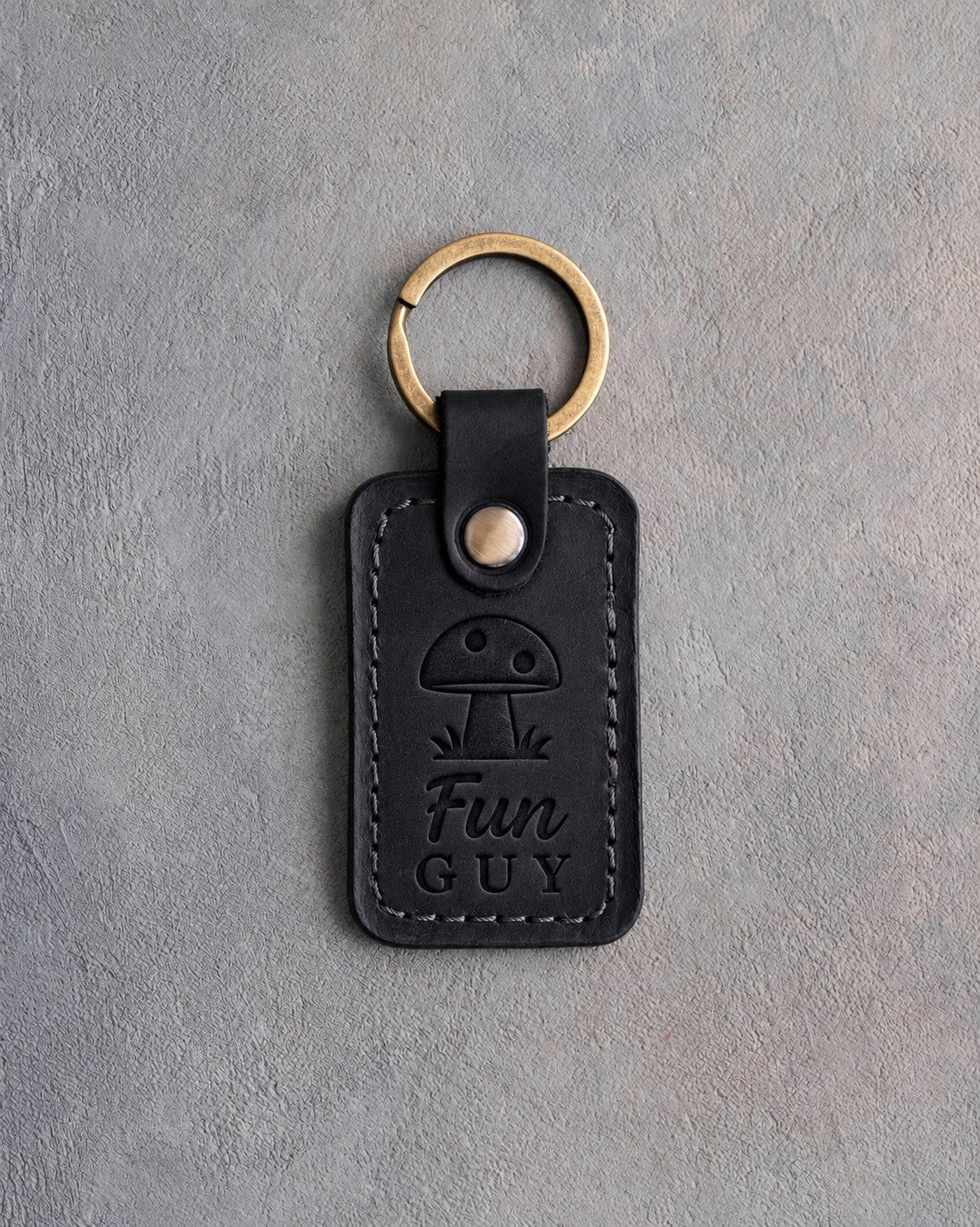 Black leather keychain debossed with a spotted mushroom above Fun GUY, on gray concrete