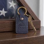Navy Daniel Gave All 2011 keychain held in an open palm beside a folded flag on a wooden table