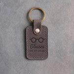 Country gray leather keychain debossed with round reading glasses above Glasses and ON MY HEAD, on gray concrete