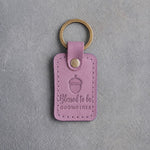 Lilac leather keychain debossed with a single acorn above Blessed to be and GODMOTHER, on gray concrete