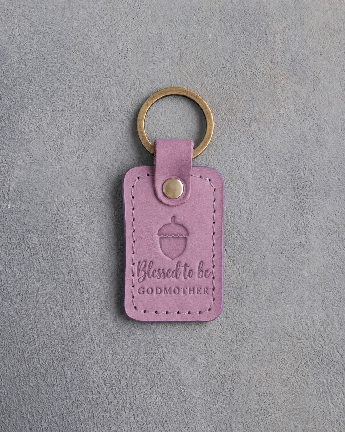 Lilac leather keychain debossed with a single acorn above Blessed to be and GODMOTHER, on gray concrete