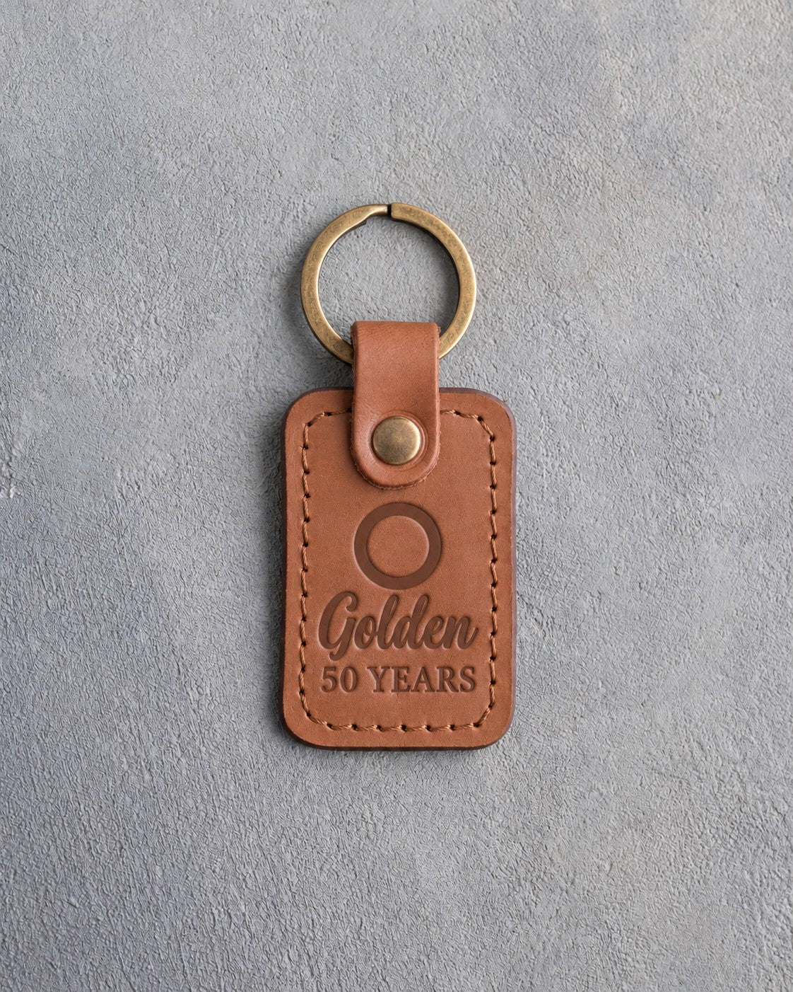 Caramel leather keychain debossed with a single wedding band above Golden and 50 YEARS, on gray concrete