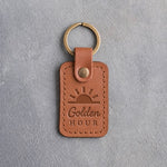 Caramel leather keychain debossed with a sun rising over the horizon above the words Golden HOUR, on gray concrete