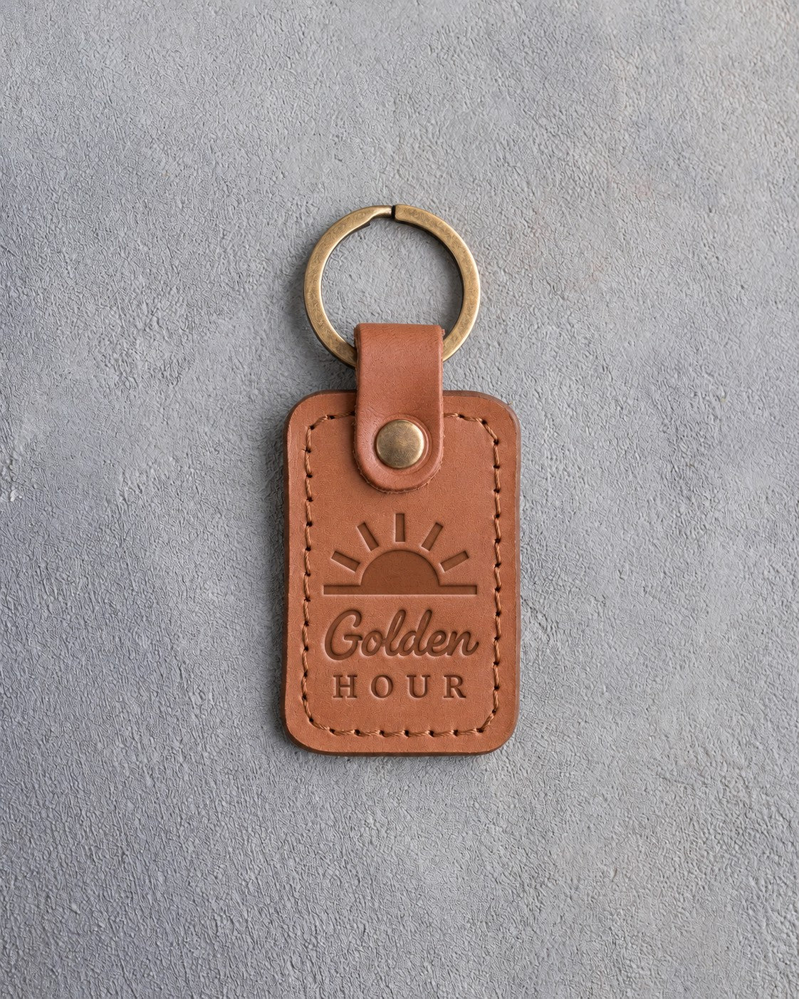 Caramel leather keychain debossed with a sun rising over the horizon above the words Golden HOUR, on gray concrete