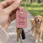 Hand holding a pink Golden Mom leather keychain in a park while a golden retriever waits on a leash