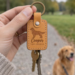 Hand holding the Cooper the Golden keychain on a park path, a golden retriever on a leash blurred behind
