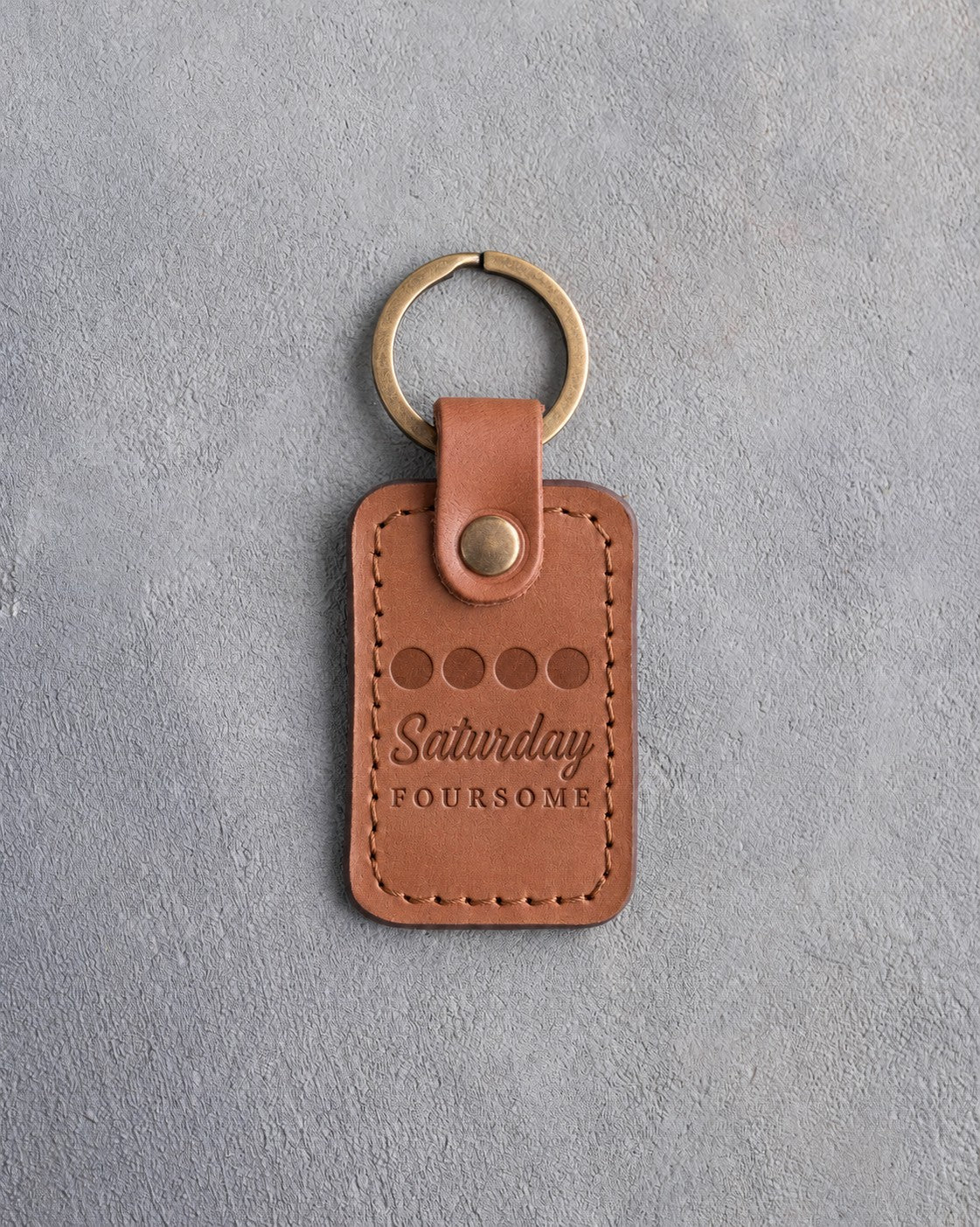 Caramel leather keychain debossed with four golf balls in a row above Saturday FOURSOME, on gray plaster