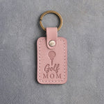 Blush pink leather keychain debossed with a golf ball on a tee above the words Golf MOM, on gray plaster