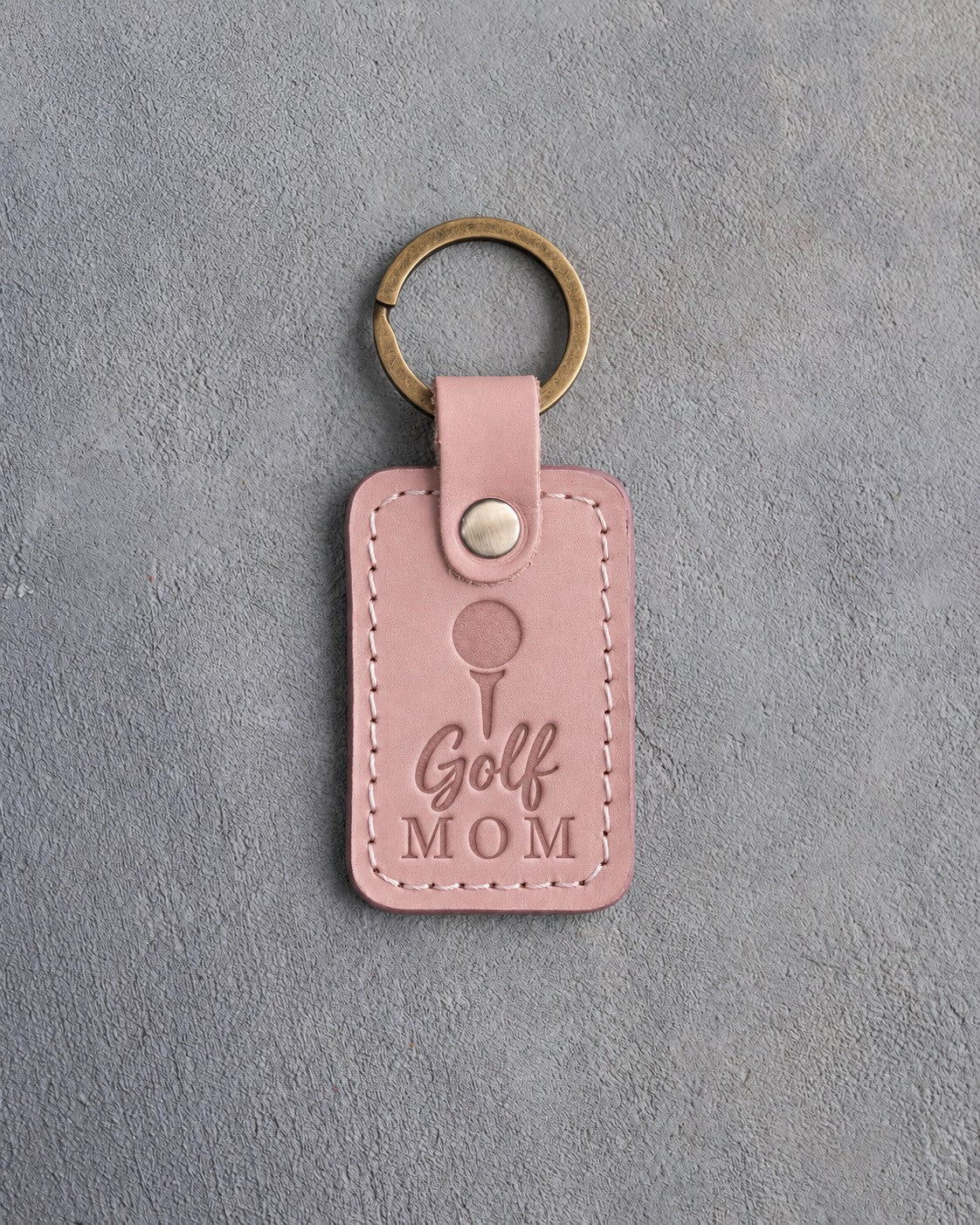 Blush pink leather keychain debossed with a golf ball on a tee above the words Golf MOM, on gray plaster