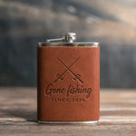 Arizona leather-wrapped hip flask debossed with crossed fishing rods above the words Gone fishing and SINCE 2026, on a wooden table