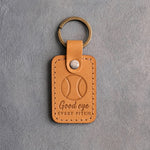 Golden brown leather keychain debossed with a baseball and the words Good eye EVERY PITCH, on gray plaster