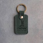 Forest green leather keychain debossed with a cowboy boot above Good hand and HARD TO FIND, on gray plaster