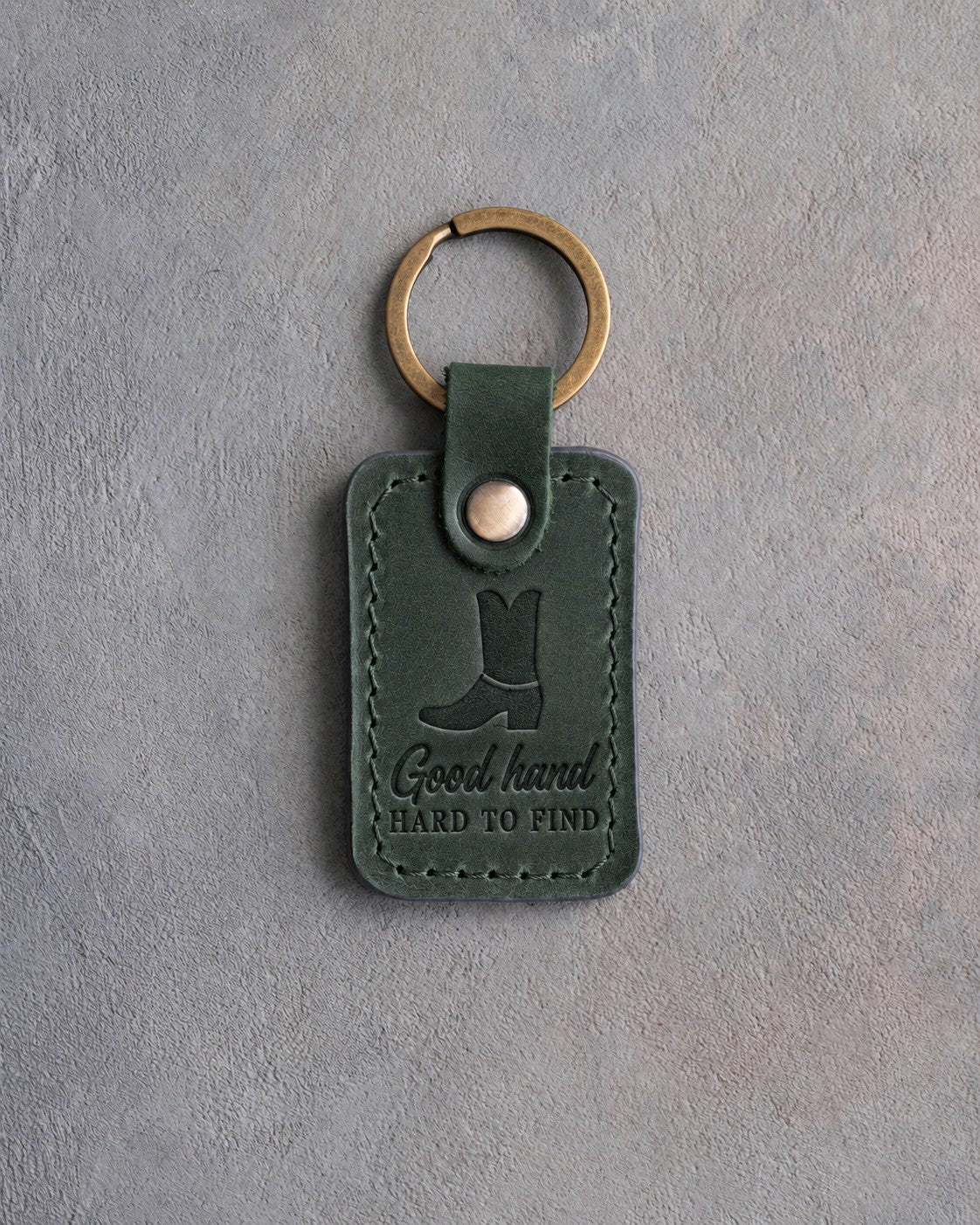 Forest green leather keychain debossed with a cowboy boot above Good hand and HARD TO FIND, on gray plaster