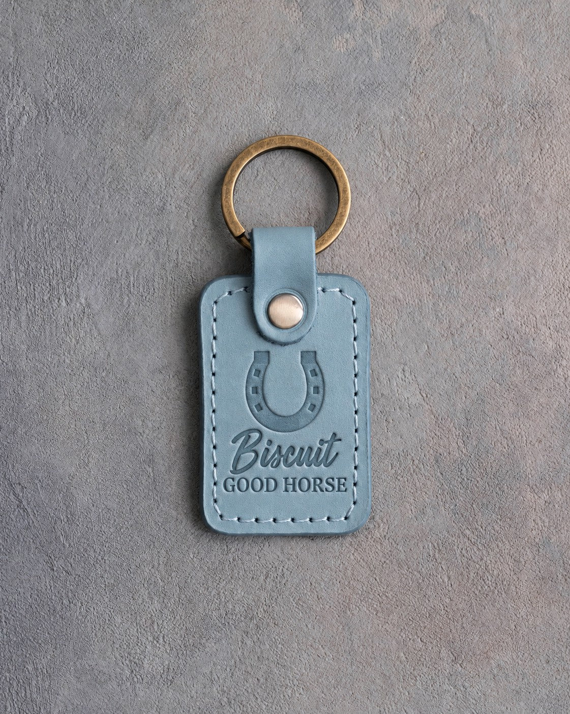 Turquoise leather keychain debossed with a heels-up horseshoe above the name Biscuit and GOOD HORSE, on gray plaster