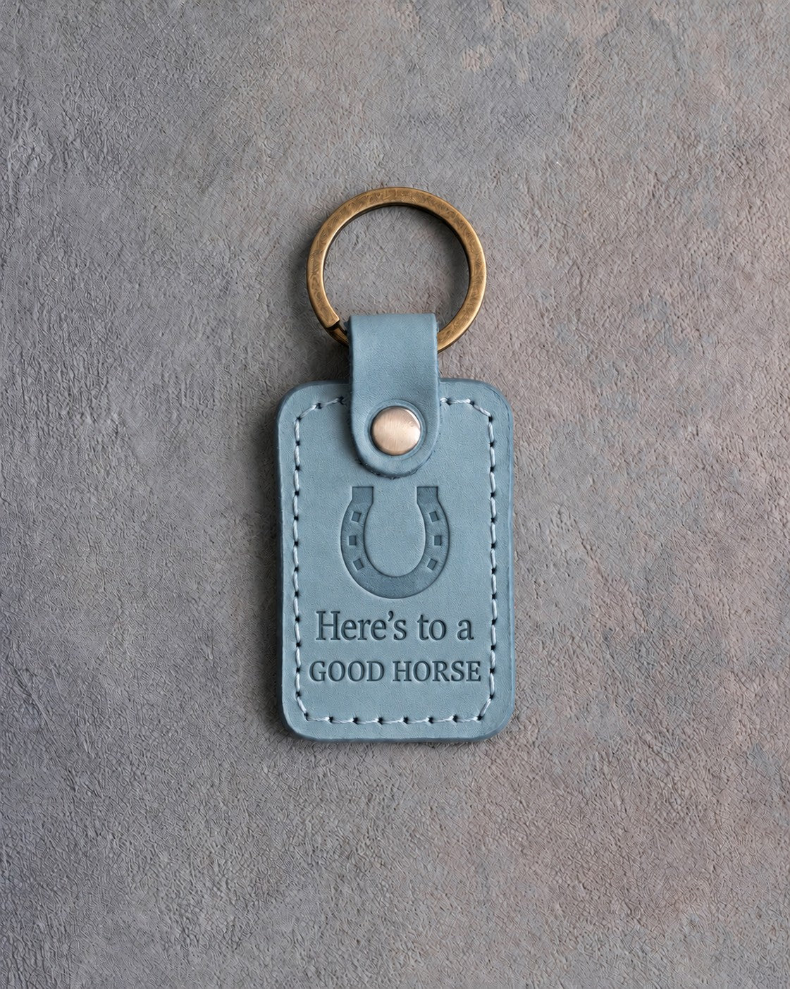 Turquoise leather keychain debossed with a heels-up horseshoe above Here's to a and GOOD HORSE, on gray concrete