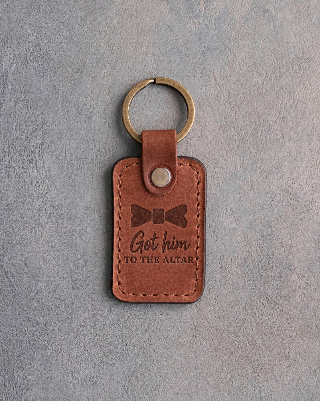 Arizona tan leather keychain debossed with a bow tie above the words Got him TO THE ALTAR, on gray concrete
