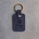 Blueberry navy leather keychain debossed with a paw print above Happy and GOTCHA DAY, on gray concrete