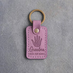 Lilac leather keychain debossed with a small hand inside a large hand above Grandma and HOLDS OUR HEARTS, on gray concrete