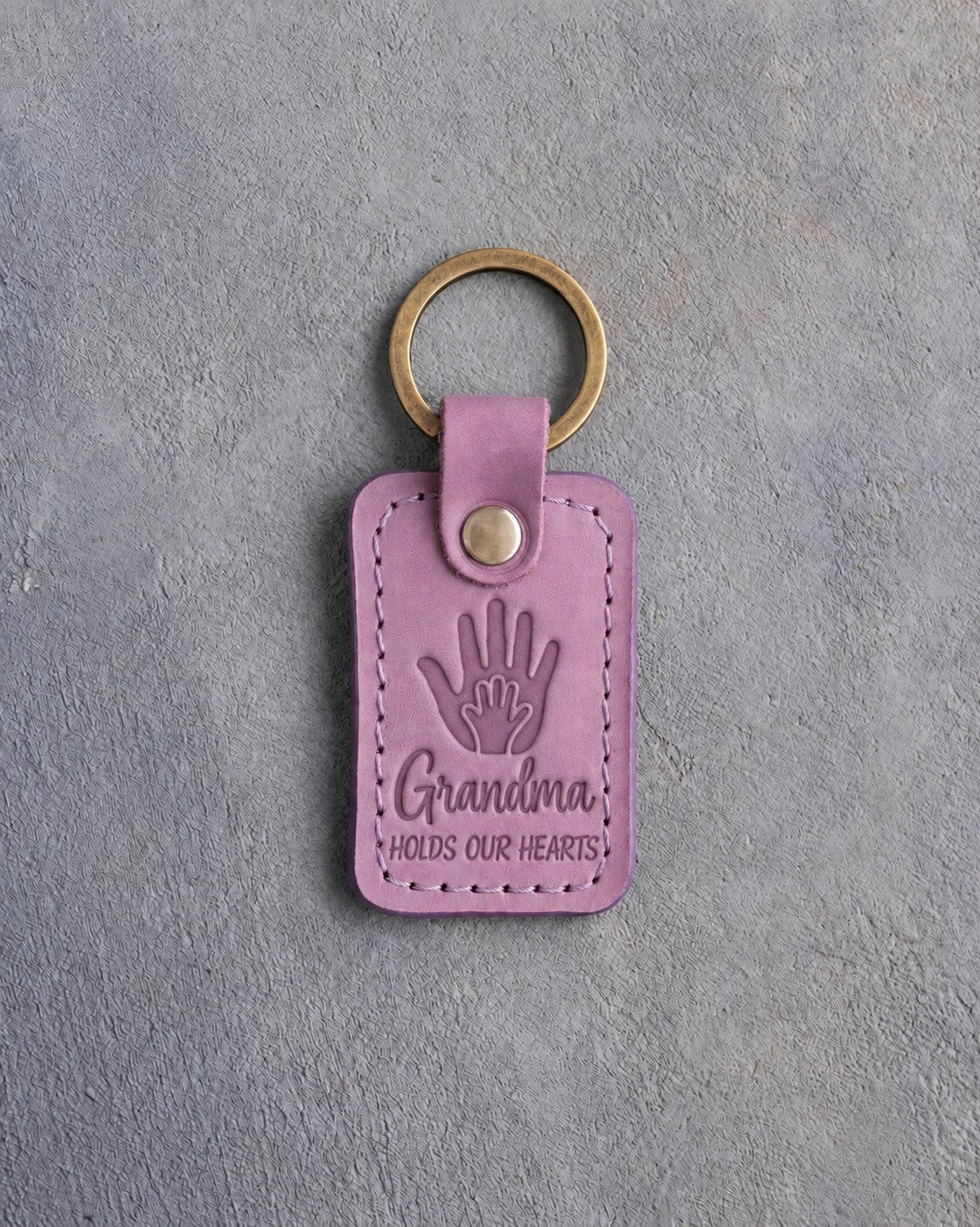 Lilac leather keychain debossed with a small hand inside a large hand above Grandma and HOLDS OUR HEARTS, on gray concrete