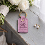 Grandma OF THE BRIDE keychain in lilac leather on a gray painted windowsill beside pearl earrings in soft light