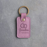 Lilac leather keychain debossed with two interlocked wedding rings above Grandma and OF THE BRIDE, on gray concrete