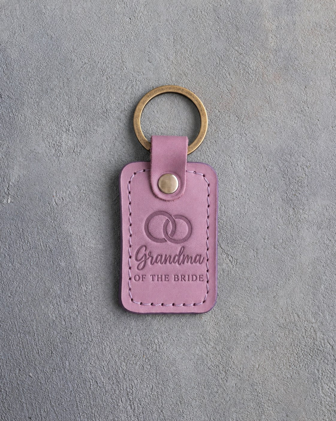 Lilac leather keychain debossed with two interlocked wedding rings above Grandma and OF THE BRIDE, on gray concrete