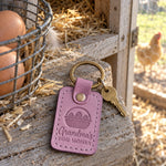 Lilac Grandma's Egg Money keychain on a weathered pine nesting box ledge beside brown eggs in a wire basket, a hen soft in the run behind