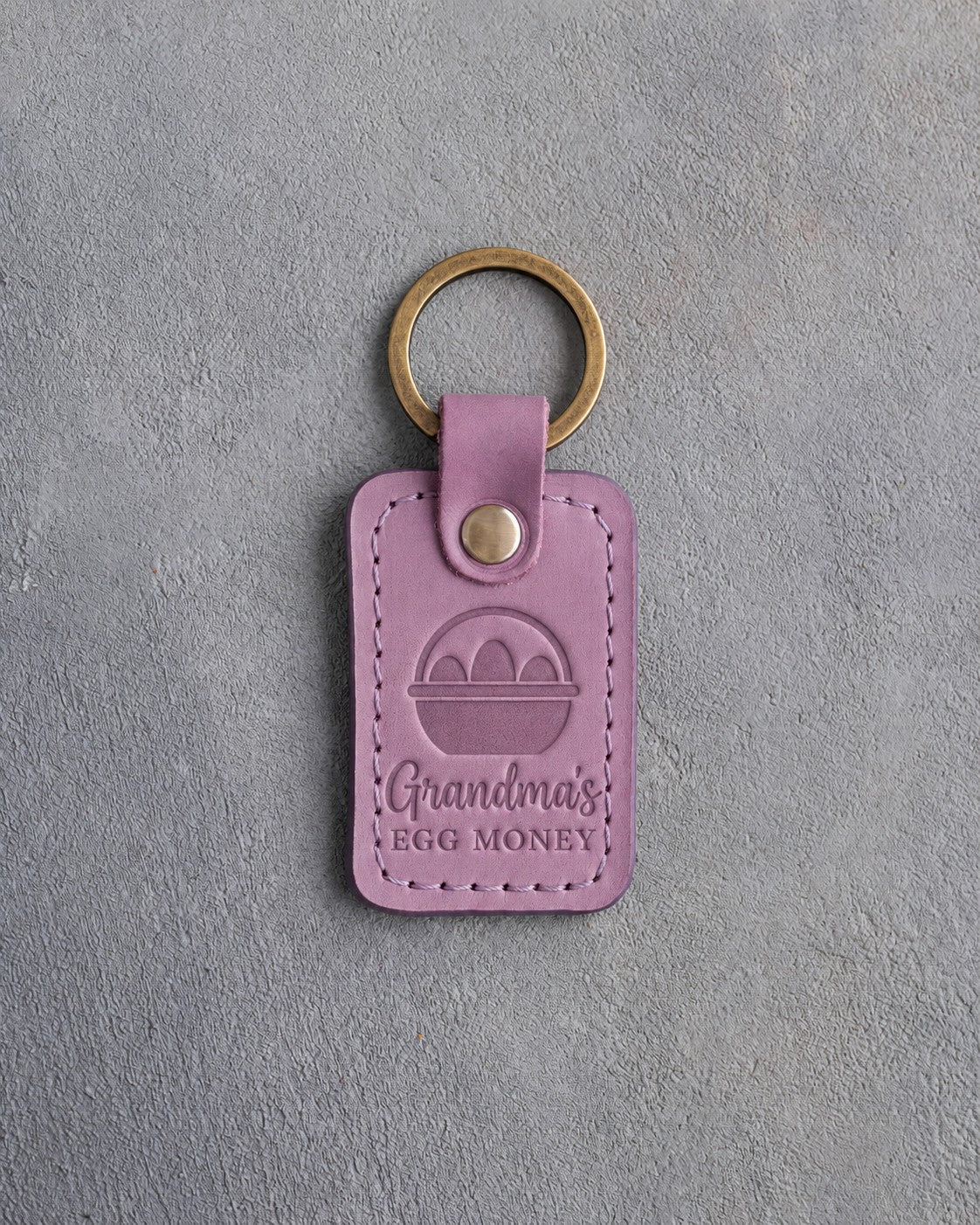 Lilac leather keychain debossed with an egg basket above the words Grandma's EGG MONEY, on gray concrete