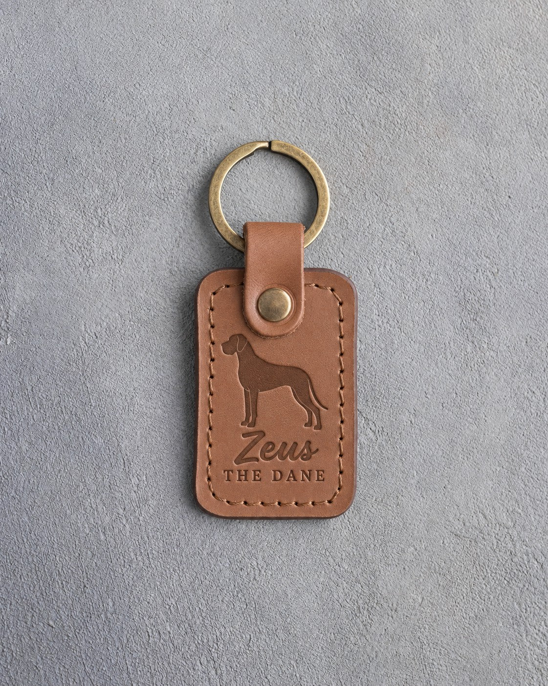 Caramel leather keychain debossed with a tall Great Dane above the name Zeus and THE DANE, on gray concrete