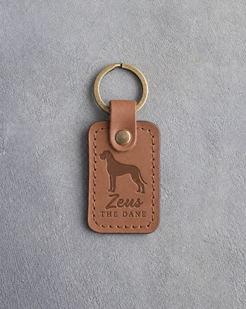 Caramel leather keychain debossed with a tall Great Dane above the name Zeus and THE DANE, on gray concrete