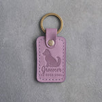 Lilac leather keychain debossed with a fluffy freshly groomed dog and the words Groomer OF GOOD DOGS, on gray concrete