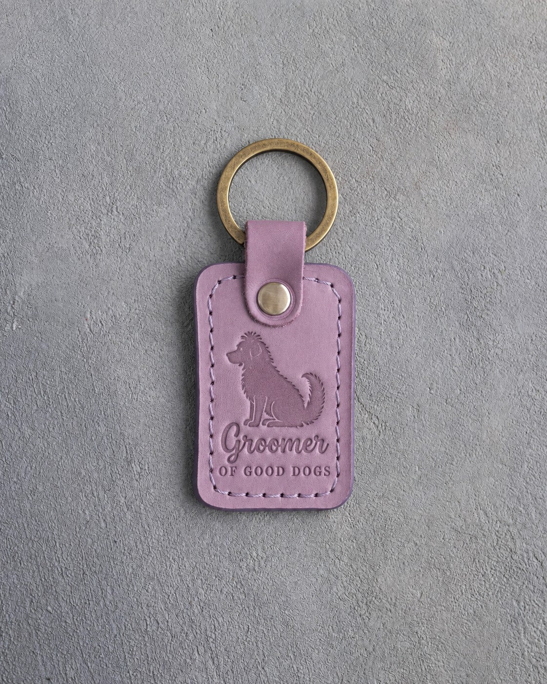Lilac leather keychain debossed with a fluffy freshly groomed dog and the words Groomer OF GOOD DOGS, on gray concrete