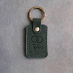 Forest green leather keychain debossed with two interlocked wedding rings above the name Tyler and GROOMSMAN, on gray concrete