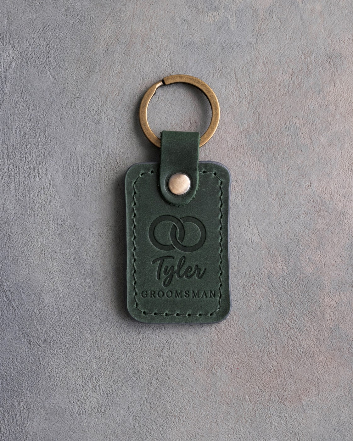 Forest green leather keychain debossed with two interlocked wedding rings above the name Tyler and GROOMSMAN, on gray concrete