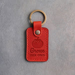Poppy red leather keychain debossed with a ripe tomato above Grows HER OWN, on gray concrete