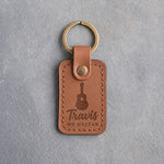 Caramel leather keychain debossed with an acoustic guitar above the name Travis and ON GUITAR, on gray plaster