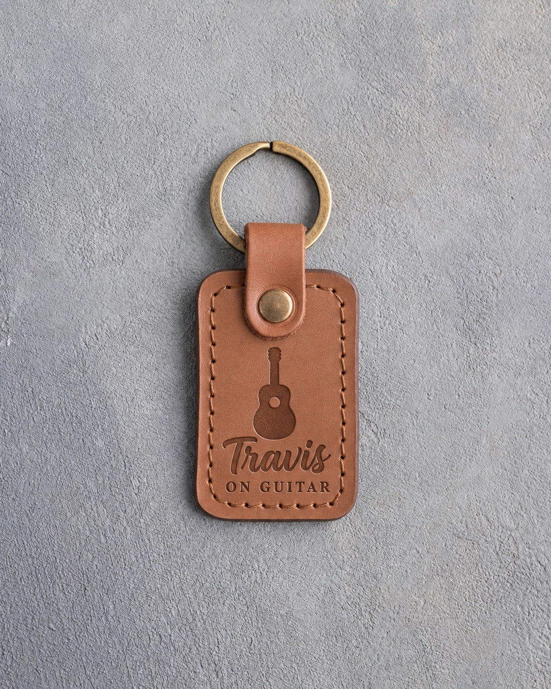 Caramel leather keychain debossed with an acoustic guitar above the name Travis and ON GUITAR, on gray plaster